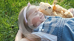 Pretty little child girl laying down with her teddy bear toy on blanket on green grass in summer smiling happily.