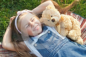 Pretty little child girl laying down with her teddy bear toy on blanket on green grass in summer smiling happily