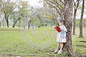 Pretty little child girl reading book in park outdoor standing lean against tree trunk in summer garden