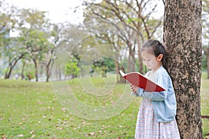 Pretty little child girl reading book in park outdoor standing lean against tree trunk in summer garden