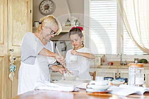 Pretty little curly girl with her happy grandmother cooking together at kitchen table.