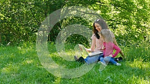 Pretty Little Daughter with Her Young Mother are Sitting on the Green Grass in the City Park with White Laptop, Looking