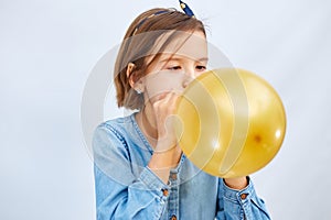 Pretty little girl in casual denim dress blowing, inflate yellow balloon