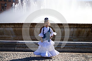 A pretty little girl dancing flamenco dressed in a white dress with ruffles and blue fringes in a famous square in seville, spain photo