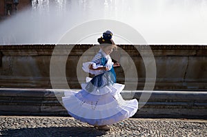 A pretty little girl dancing flamenco dressed in a white dress with ruffles and blue fringes in a famous square in seville, spain photo