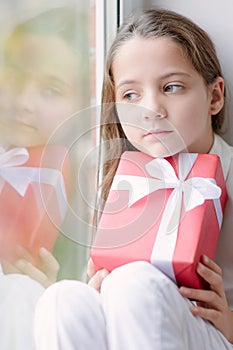 Pretty little girl dreaming by the window holding gift