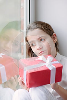 Pretty little girl dreaming by the window holding gift