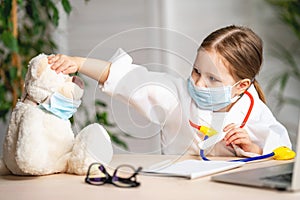 Pretty little girl dressed in a white lab coat and mask was taking the temperature of a Teddy bear