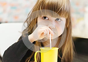 A pretty little girl drinks milk with a drinking straw from a yellow plastic mug. A little girl is sitting at the table and having