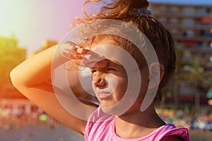 A pretty little girl looks far away from right to left side, squinting and playing the ape in sunshine on city beach background. S