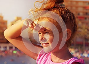 A pretty little girl looks to far away from right to left side, smiling and squinting in sunshine on city beach background. Sunset