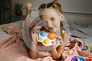 Pretty little girl in pink pajamas laying in her bad at daytime and playing antistress multicolored toy popit. Trendy antistress t