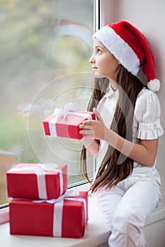 Pretty little girl in Santa hat dreaming by the window holding gift