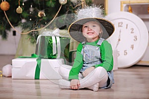 Pretty little girl in shine hat smiling with present near the Christmas tree