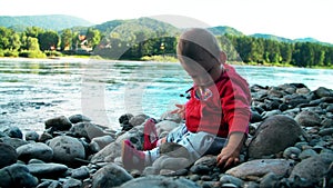 Pretty little girl is sitting on a bank of mountain river and playing with stones.