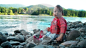 Pretty little girl is sitting on a bank of mountain river and playing with stones.