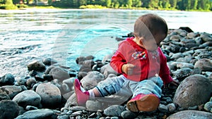 Pretty little girl is sitting on a bank of mountain river and playing with stones.