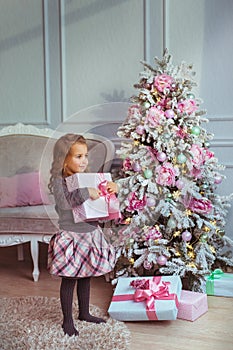 Pretty little girl stands with a gift box at hands near Christmas tree and looks to side