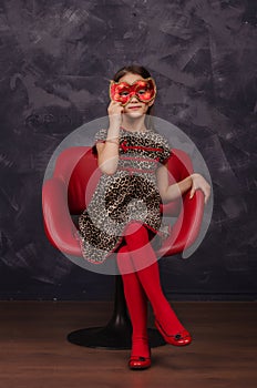 Pretty little girl wearing beautiful dress sitting in red armchair. She is wearing red masquerade carnival mask. Studio shot.