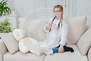 Pretty little girl wearing doctor coat, sitting on the sofa with legs curled up, ponting finger up, looking at the photo