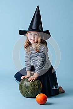 Pretty little girl in witch hat playing with green pumpkin, leaning on it