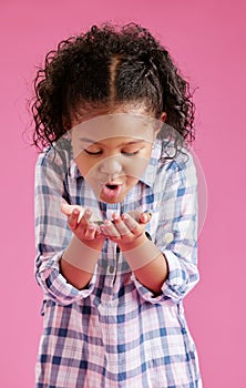 A pretty little mixed race girl with curly hair blowing on coins in her hands against a pink copyspace background in a