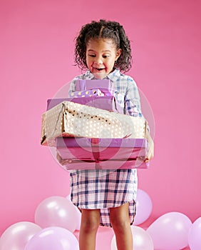 A pretty little mixed race girl with curly hair holding a stack of wrapped presents against a pink copyspace background