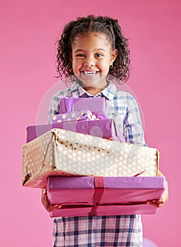 A pretty little mixed race girl with curly hair holding a stack of wrapped presents against a pink copyspace background