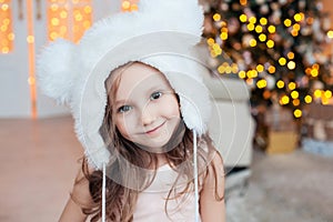 Pretty little smiling girl wearing white fur hat posing against Christmas tree indoors.