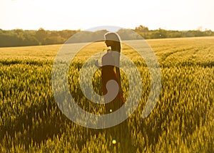 Pretty long haired slim woman in long dress enjoys the sunset next to a meadow, and holds a piece of crop in hand.