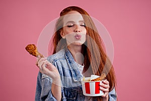 Pretty long redhead ginger girl eating fries chicken in studio pink background