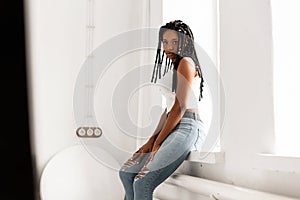 Pretty lovely African American woman with dreadlocks in white fashion youth top in blue stylish jeans relaxÐµs near window in