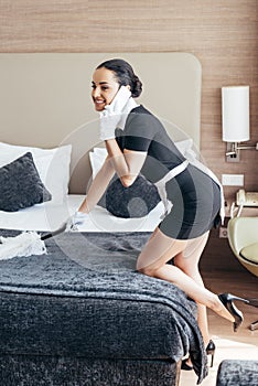 Pretty maid in white gloves cleaning bed with duster and talking on smartphone in hotel room