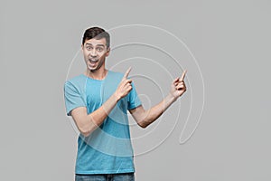 Pretty man in a blue tee pointing with a finger at the copy space isolater over grey background.