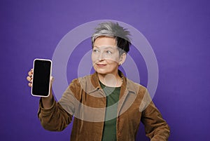 Pretty mature woman showing smartphone blank screen advertising mobile application posing isolated over violet studio background.