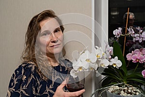 A pretty middle-aged woman 45 years old stands at the window with flowers with a glass of red holiday wine