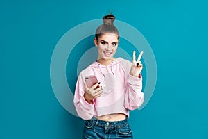 Pretty millennial hipster girl holding mobile phone and doing victory sign