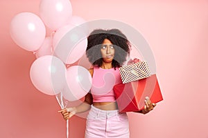 Pretty mixed race woman, holding pink balloons and presents given for her birthday , looks a side with bored look