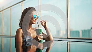 Pretty mixed race woman in sunglasses and bikini relaxing in swimming pool and drinking cocktail. Gorgeous lady enjoying