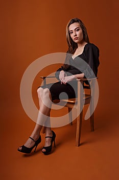 Pretty model in black dress, highheel shoes sits in chair on orange background