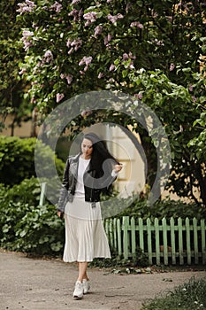 Pretty model girl in summer midi skirt and leather jacket posing near a bloomy tree. Young brunette woman in spring