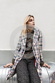 Pretty modern young woman blonde in checkered stylish blazer in fashionable pants sits on metal pipe near vintage building on