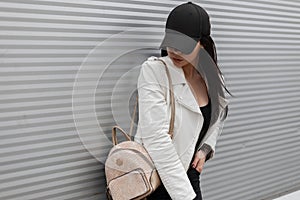 Pretty modern young woman in a fashionable black baseball cap in a vintage white leather jacket in jeans with a gold backpack