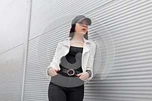 Pretty modern young woman in a white leather jacket in vintage black jeans in a stylish baseball cap is resting standing