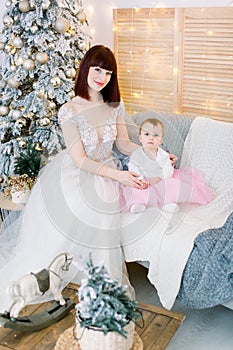 Pretty mother with her baby girl, wearing in fashionable elegant dresses, sitting on stylish gray sofa on the background