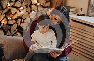 Pretty mother and her little daughter reading book in the evening at home with cozy and christmas decoration. Beautiful woman and