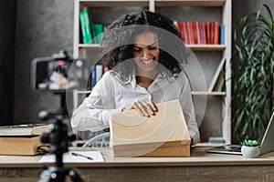 Pretty multinational woman in casual wear recording video on camera while unpacking gift box.