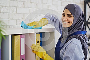 Pretty Muslim woman cleaning bookshelves with a rug in the office