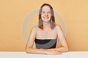 Pretty pleased Caucasian female model wearing black top sitting at table isolated over beige background looking at camera with