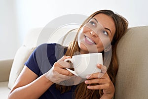 Pretty positive dreamy girl holds cup with warm beverage imagining sitting on sofa